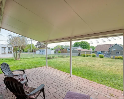 Covered patio with brick pavers and three black plastic chairs facing a green backyard with a chain-link fence and neighboring houses in the distance.