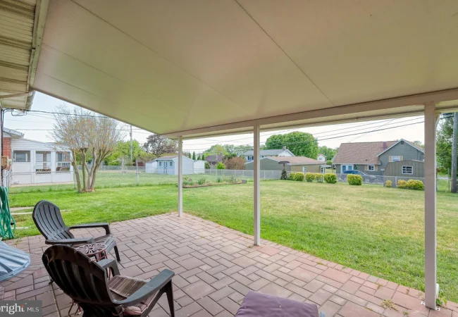 Covered patio with brick pavers and three black plastic chairs facing a green backyard with a chain-link fence and neighboring houses in the distance.