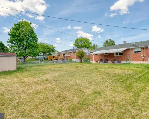 Spacious backyard with a brick house, covered patio, and a wooden playset; a shed sits near a chain-link fence under a bright sky.