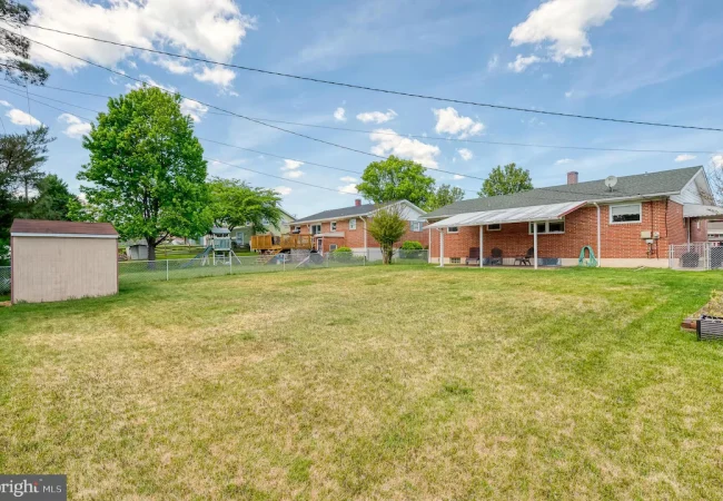 Spacious backyard with a brick house, covered patio, and a wooden playset; a shed sits near a chain-link fence under a bright sky.