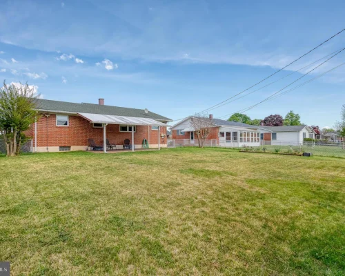 Backyard of a single-story brick house with a covered patio, outdoor seating, and a lawn bordered by a chain-link fence under a blue sky