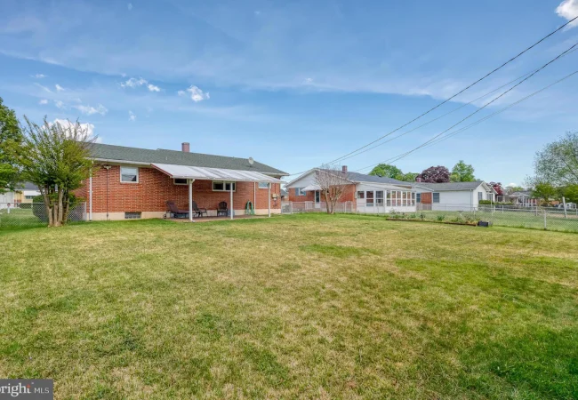 Backyard of a single-story brick house with a covered patio, outdoor seating, and a lawn bordered by a chain-link fence under a blue sky