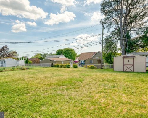 Suburban backyard with a beige shed, chain-link fence, and trimmed lawn, facing neighboring houses and utility poles under a blue, partly cloudy sky