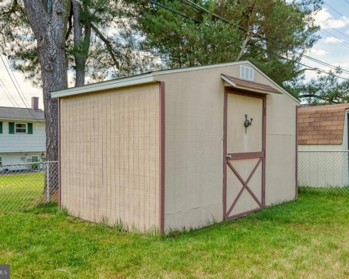 Beige storage shed with brown trim and a cross-braced door in a fenced residential backyard, with a large tree and neighboring houses in the background.