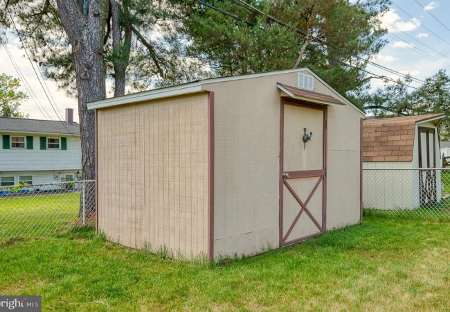 Beige storage shed with brown trim and a cross-braced door in a fenced residential backyard, with a large tree and neighboring houses in the background.
