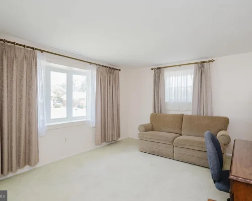 Living room featuring a beige sofa, a wooden desk with a chair, and large windows with heavy drapes.