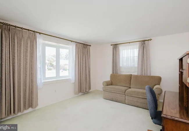 Living room featuring a beige sofa, a wooden desk with a chair, and large windows with heavy drapes.