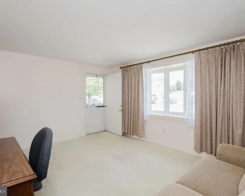 Living room with a beige sofa, wooden desk and chair, white walls, and large window with sheer and beige curtains.