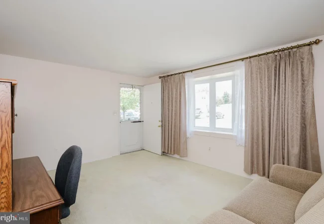 Living room with a beige sofa, wooden desk and chair, white walls, and large window with sheer and beige curtains.