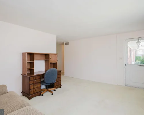 Wooden desk with built-in shelves and a blue rolling office chair in a sparsely furnished living room with light walls and a door outside.