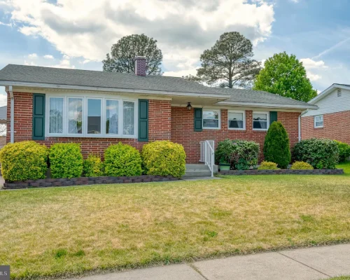 Single-story brick house with green shutters and a manicured front garden of shrubs and small trees.