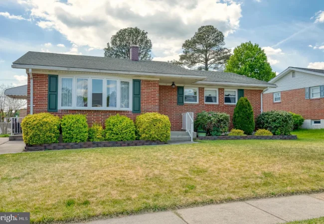 Single-story brick house with green shutters and a manicured front garden of shrubs and small trees.