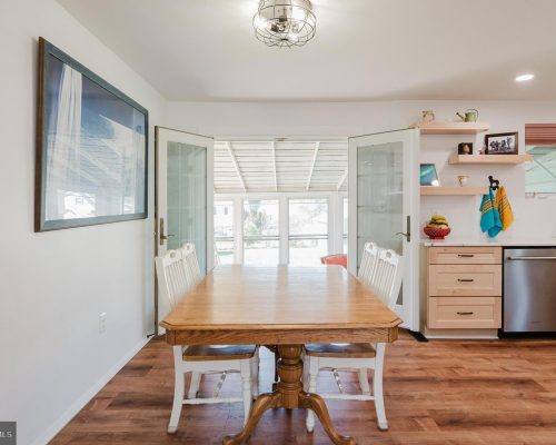 Bright dining area with a wooden table and white chairs, open French doors leading to a sunlit room beyond.