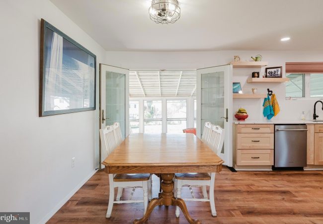 Bright dining area with a wooden table and white chairs, open French doors leading to a sunlit room beyond.
