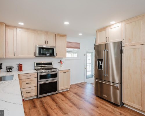 Bright kitchen with light wood cabinets, stainless steel fridge, and double ovens against a white wall. Sturdy hardwood floors run through the space.] ,
