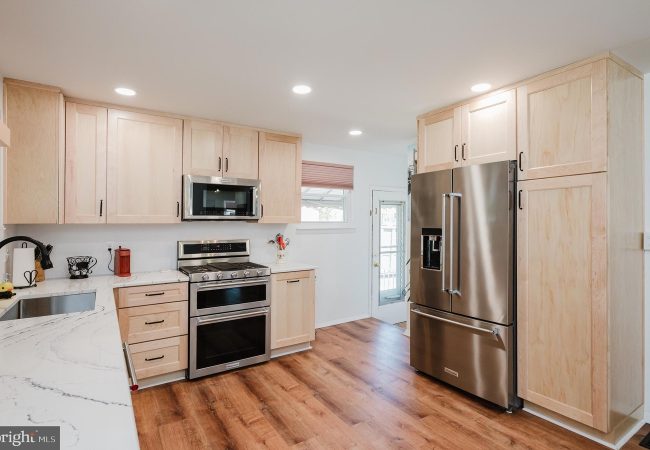 Bright kitchen with light wood cabinets, stainless steel fridge, and double ovens against a white wall. Sturdy hardwood floors run through the space.] ,