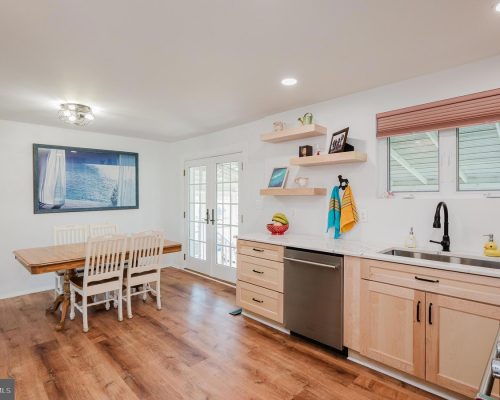 Bright kitchen and dining area with a wooden dining table and white chairs, light wood cabinets, and a stainless dishwasher beneath a marble-like counter.