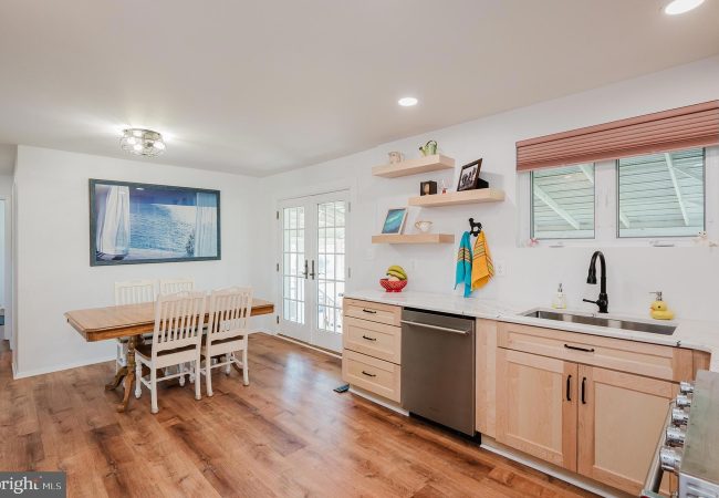 Bright kitchen and dining area with a wooden dining table and white chairs, light wood cabinets, and a stainless dishwasher beneath a marble-like counter.