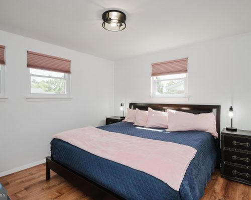 Bedroom with a dark wood bed, navy quilt, and pale pink pillows; pink throw at foot and matching blinds in three windows.