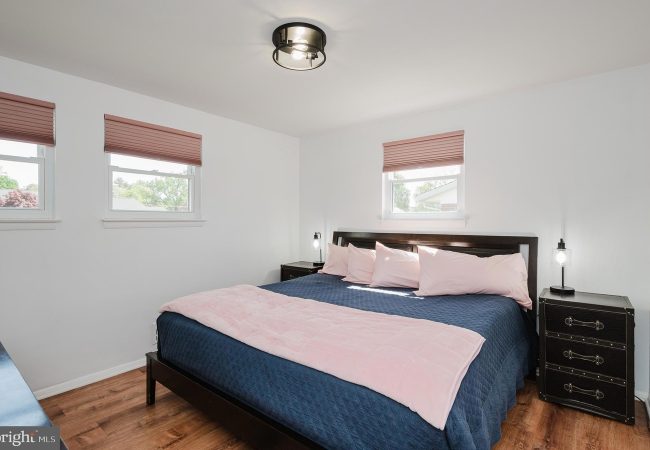 Bedroom with a dark wood bed, navy quilt, and pale pink pillows; pink throw at foot and matching blinds in three windows.