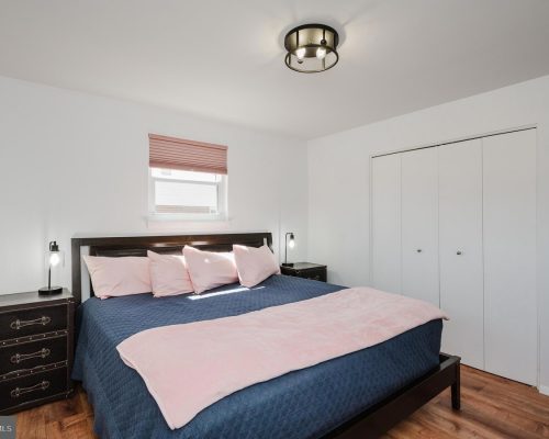 Bedroom with a dark wood bed, navy quilt, pink pillows and throw, two matching nightstands with lamps, small window with a pink shade, and white double closet doors.