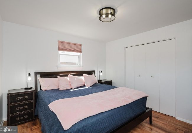 Bedroom with a dark wood bed, navy quilt, pink pillows and throw, two matching nightstands with lamps, small window with a pink shade, and white double closet doors.