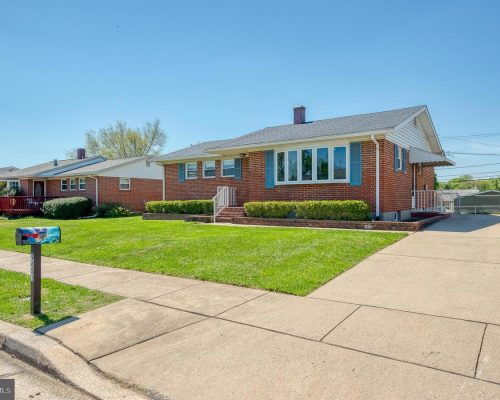 Brick ranch-style house with a manicured front lawn, hedges, and a long driveway on the right under a clear blue sky.