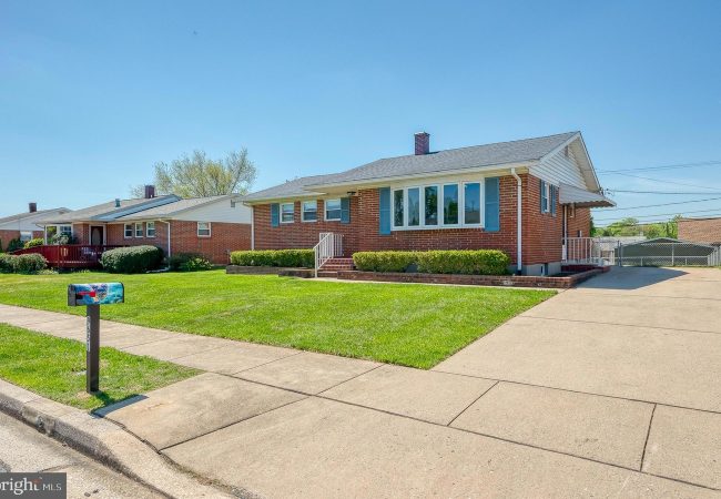 Brick ranch-style house with a manicured front lawn, hedges, and a long driveway on the right under a clear blue sky.