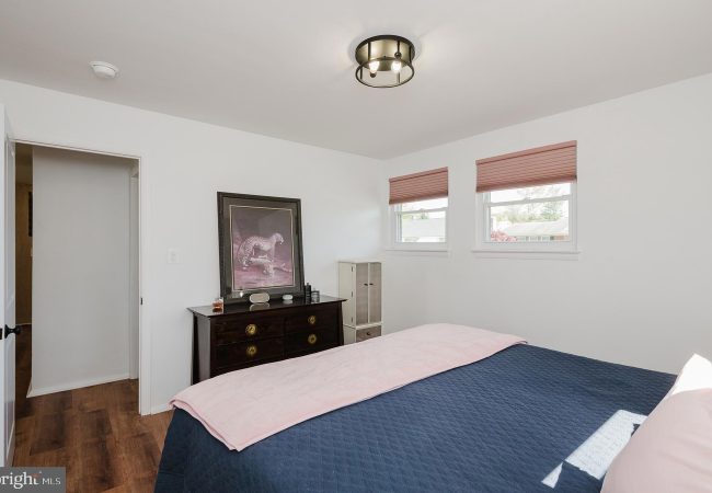 Bedroom with white walls, a dark dresser topped by a framed leopard print, and a bed covered in a navy quilt with a pink throw. Limit: concise and informative for screen readers.