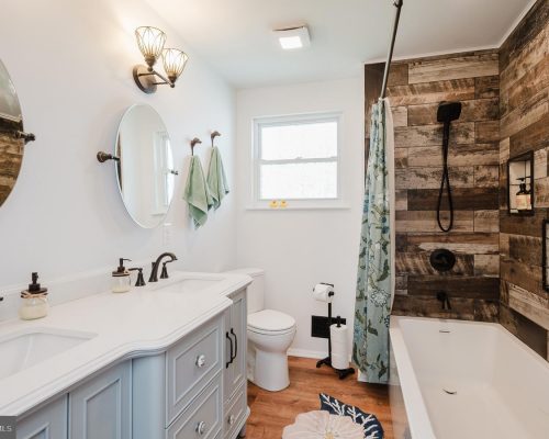 Bright bathroom with a light blue double-sink vanity, two round mirrors, and black fixtures next to a bathtub/shower combo with wood-paneled walls and a blue shower curtain