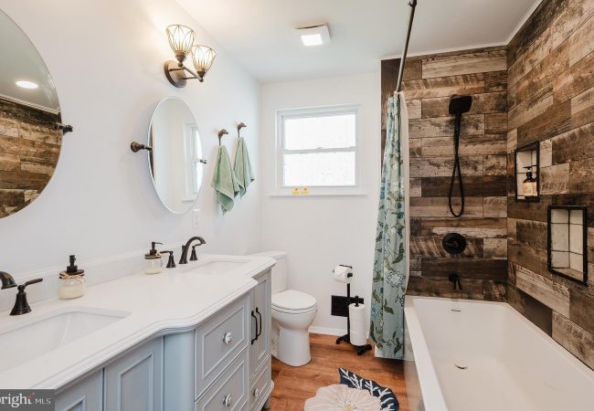 Bright bathroom with a light blue double-sink vanity, two round mirrors, and black fixtures next to a bathtub/shower combo with wood-paneled walls and a blue shower curtain