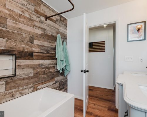 Bathroom with white tub and rustic wood plank wall; teal towels on hooks, and a curved white vanity with a sink nearby.