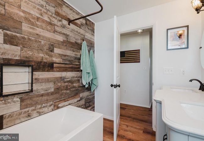 Bathroom with white tub and rustic wood plank wall; teal towels on hooks, and a curved white vanity with a sink nearby.