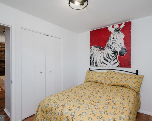 Small bedroom with white walls and a twin bed in a yellow floral quilt, black metal headboard, and a bold red zebra painting above the bed.
