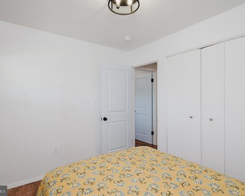 Bright, minimalist bedroom with white walls, a bed in the foreground covered by a yellow floral quilt, and white closet doors along one wall; a doorway leads to another room; small window on the left side.