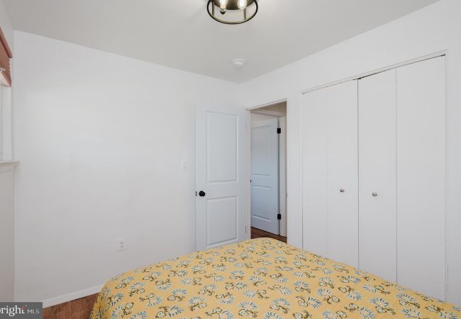 Bright, minimalist bedroom with white walls, a bed in the foreground covered by a yellow floral quilt, and white closet doors along one wall; a doorway leads to another room; small window on the left side.