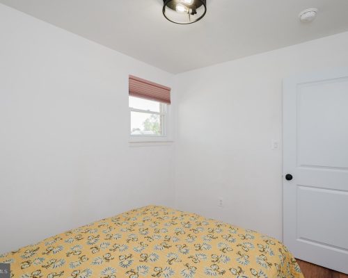 Small white bedroom with a bed covered in a yellow floral quilt, a window with pink blinds, and a white paneled door to the right. A ceiling light hangs above.