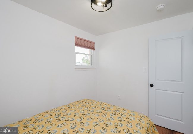 Small white bedroom with a bed covered in a yellow floral quilt, a window with pink blinds, and a white paneled door to the right. A ceiling light hangs above.