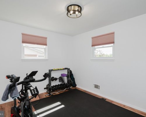 Small home gym in a bright white room featuring a stationary bike, a dumbbell rack with weights, a black exercise mat, and a few yoga accessories by the wall beneath two pink-blind windows.
