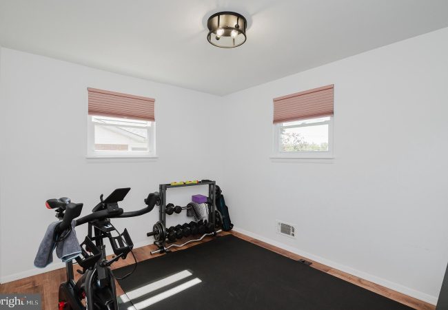 Small home gym in a bright white room featuring a stationary bike, a dumbbell rack with weights, a black exercise mat, and a few yoga accessories by the wall beneath two pink-blind windows.