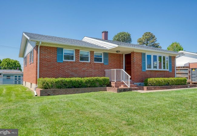 Front view of a single-story brick ranch-style house with blue shutters, white railing, and a brick staircase leading to the front door, surrounded by a green lawn and trimmed hedges.