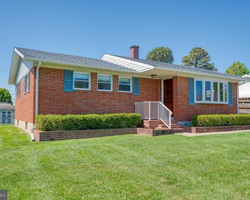 Brick single-story house with blue shutters and a white railing at the front steps, set in a green lawn under a clear blue sky.