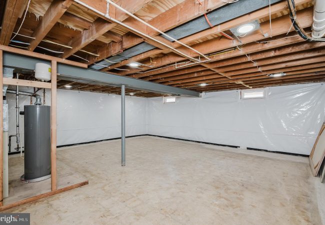 Unfinished basement with exposed joists, a grey water heater, a vertical support post, and white plastic-wrapped walls.
