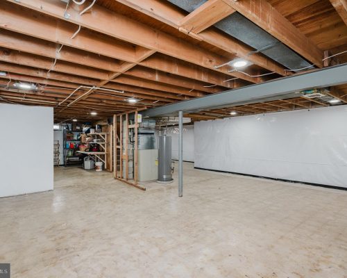 Basement with exposed wooden ceiling beams, a storage area, and a water heater against a white wall. sharp concrete floor.