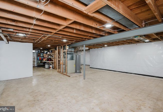 Basement with exposed wooden ceiling beams, a storage area, and a water heater against a white wall. sharp concrete floor.