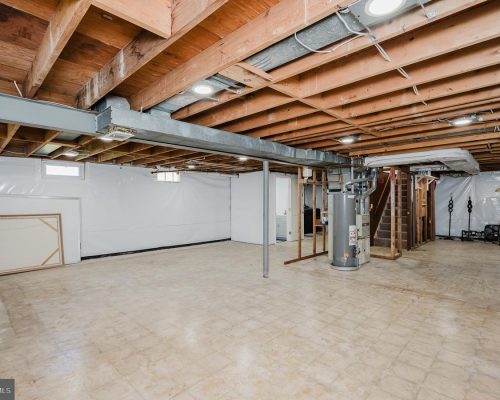 Unfinished basement with exposed wooden joists, a central furnace, and utility area along the back wall.