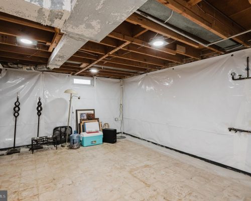 Unfinished basement with plastic-walled sides, exposed ceiling beams, and stored items like a cooler, water jug, lamp, and framed pictures.]','Basement corner with a floor lamp, cooler, water jug, and stacked framed artwork against plastic walls.','Utility basement scene showing exposed pipes on the right and various storage items scattered around.','Cluttered basement storage area with a standing lamp, cooler, water jug, and several frames propped against the wall.','A spacious basement room with white plastic sheeting on the walls, concrete floor, and miscellaneous belongings.