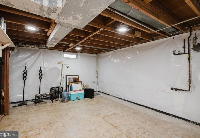 Unfinished basement with plastic-walled sides, exposed ceiling beams, and stored items like a cooler, water jug, lamp, and framed pictures.]','Basement corner with a floor lamp, cooler, water jug, and stacked framed artwork against plastic walls.','Utility basement scene showing exposed pipes on the right and various storage items scattered around.','Cluttered basement storage area with a standing lamp, cooler, water jug, and several frames propped against the wall.','A spacious basement room with white plastic sheeting on the walls, concrete floor, and miscellaneous belongings.