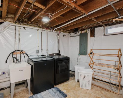Basement laundry area with a washer and dryer, utility sink, laundry basket, and a wooden drying rack against white plastic-wrapped walls.