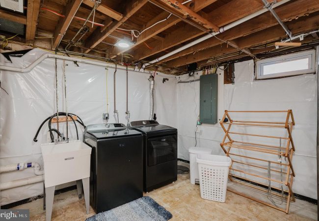 Basement laundry area with a washer and dryer, utility sink, laundry basket, and a wooden drying rack against white plastic-wrapped walls.